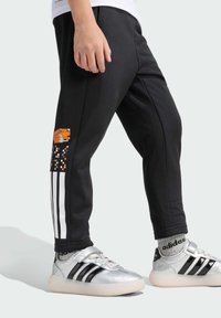 Černé tepláky s bílými pruhy, zdobené oranžovou pixelovanou grafikou. V kombinaci se stříbrno-černými teniskami a šedými ponožkami.