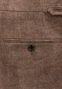 Tela marrón con un tejido texturizado. Presenta un botón negro único y un pequeño bolsillo con una línea de costura encima.