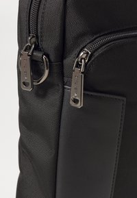 Μαύρο σακίδιο νάιλον με υφή, ασημένιες φερμουάρ με λογότυπο και λεία δερμάτινη λεπτομέρεια. Περιλαμβάνει διπλούς χώρους με φερμουάρ.