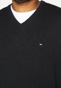 Pull en tricot noir avec un col en V. Présente un petit logo rouge, blanc et bleu sur le côté gauche de la poitrine. Texture douce et coupe ajustée.