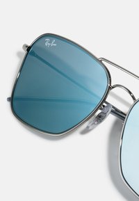 Ochelari de soare Ray-Ban cu lentile de nuanță albastră, rame metalice argintii și tampoane pentru nas reglabile. Formă pătrată a lentilelor, design minimalist.
