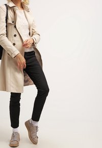 Béžový trenčkot přes bílý chlupatý svetr, černé slim-fit kalhoty a šedé tenisky s texturovanými detaily a bílou podrážkou.