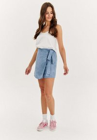 Světle modrá zavinovací sukně s detailem vázání, vyrobená z jemného materiálu, v kombinaci s bílým topem a růžovými teniskami. Hladká textura a ležérní design.