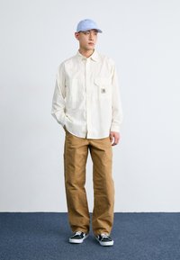 Camicia chiara, con bottoni, dotata di due tasche frontali, abbinata a pantaloni cargo beige. Il modello indossa un cappellino azzurro chiaro e sneakers nere.