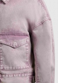 Veste en denim violet clair avec une poche à rabat sur le devant, des coutures apparentes et un col, sur un fond uni.