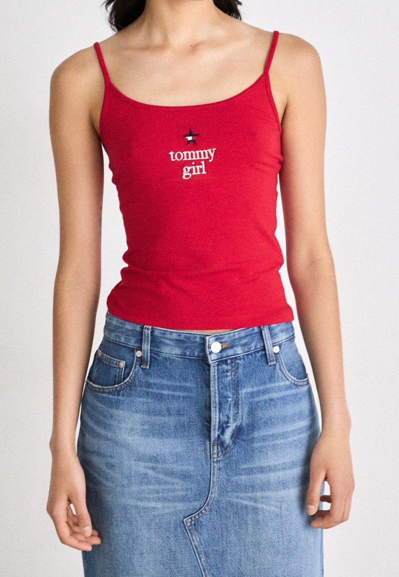 Femme portant un débardeur rouge avec le texte "tommy girl" et un logo étoile, associé à une jupe en jean bleu taille haute.