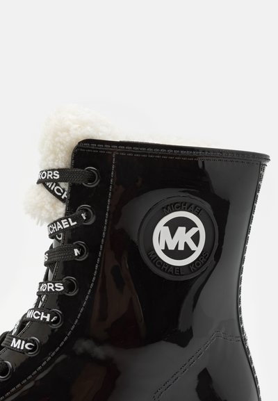 Bottines en cuir verni noir avec doublure en fausse fourrure blanche, présentant un détail de logo et des lacets noirs avec le texte "MICHAEL KORS". Design à bout rond.