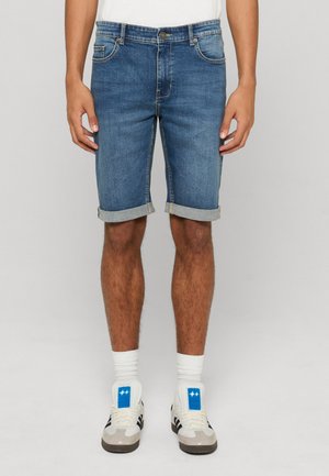 Homme portant un short en jean bleu retroussé, des chaussettes blanches et des baskets blanches avec des détails noirs et bleus, debout devant un fond uni.