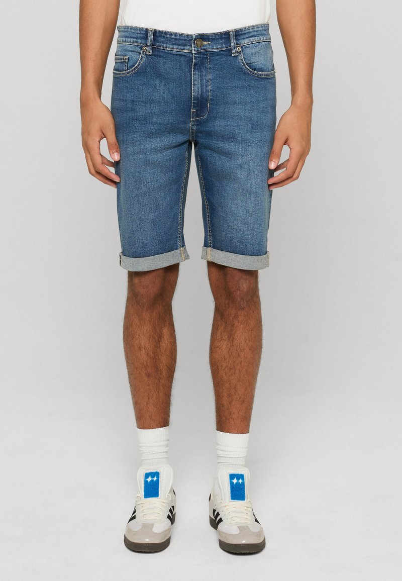 Homme portant un short en jean bleu retroussé, des chaussettes blanches et des baskets blanches avec des détails noirs et bleus, debout devant un fond uni.