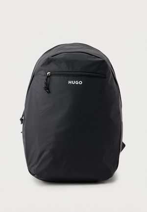 Rucsac negru HUGO cu un buzunar frontal cu fermoar, design minimalist și vârf ușor rotunjit, pe un fundal simplu gri deschis.