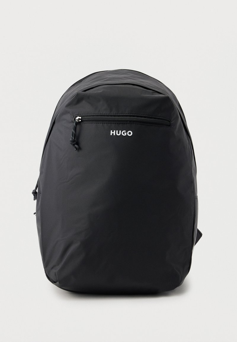 Sac à dos HUGO noir avec une poche zippée à l'avant, design minimaliste et sommet légèrement arrondi, sur un fond uni gris clair.
