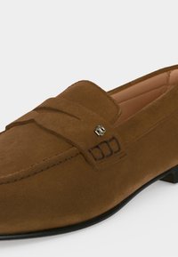 Mocassin en daim marron avec semelle noire, détails cousus et petit emblème décoratif métallique sur la bride supérieure.