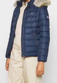 Tmavě modrá péřová bunda s kapucí lemovanou kožíškem, se zapínáním na zip vpředu a bočními kapsami. Má prošívanou strukturu a logo značky na rukávu.