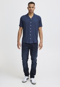 Camicia a maniche corte color navy in tessuto morbido con colletto, chiusura a bottoni, abbinata a jeans blu scuro e sneakers bianche con dettagli neri.