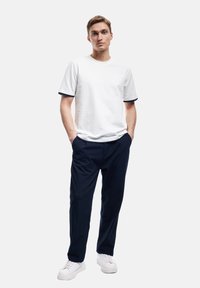 Witte T-shirt met korte mouwen en marineblauwe bies, gecombineerd met donkerblauwe broek. Model draagt witte sneakers en staat met de handen in de zakken.