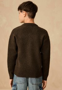 Brązowy sweter o teksturowanej powierzchni z okrągłym dekoltem i prążkowanymi mankietami, w połączeniu z jasnoniebieskimi spodniami cargo. Tkanina ma subtelny wzór.