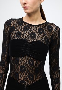 Black lace bodysuit with long sleeves, featuring a ruched, velvet bandeau top. Floral lace pattern adds texture and design detail.