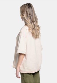 Lys beige oversized t-skjorte med korte ermer og rå kanter, parret med olivengrønn cargo bukse, sett bakfra.