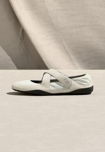 Λευκό επίπεδο παπούτσι με στρογγυλεμένη μύτη, ελαστική πίσω πλευρά και ρυθμιζόμενο λουράκι με κουμπί κλεισίματος σε μπεζ επιφάνεια.