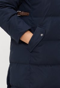 Tumedat sinise puffer-jakk, millel on tikandite tekstuur, bruna nahkrihmaga aksent ja nööbiga sulgemine mansetil.