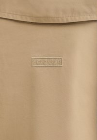 Beiger Stoff mit glatter Textur und einem eingenähten rechteckigen Aufnäher mit der Aufschrift "PCLO CLUB" in heller Stickerei.