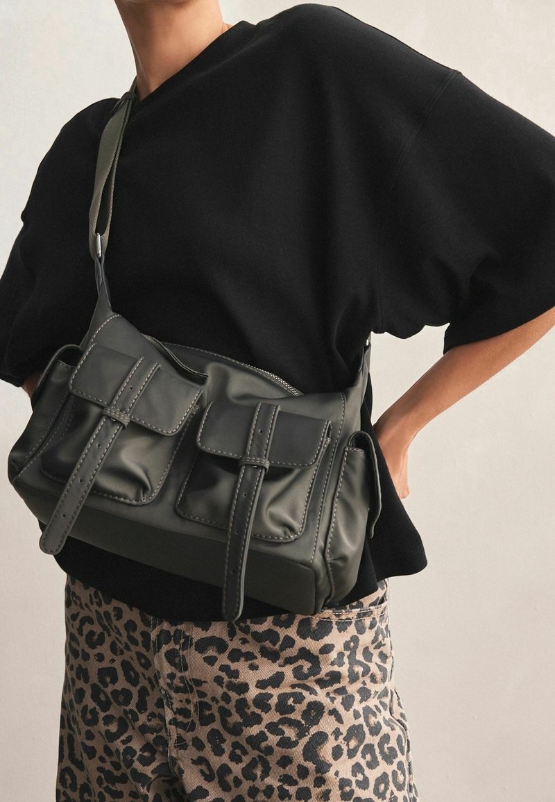 Personne portant un haut noir oversize et un pantalon à imprimé léopard avec un sac bandoulière gris foncé avec deux poches avant.