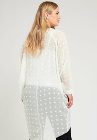 Blusa blanca de manga larga y transparente con un patrón de lunares texturizados y un dobladillo asimétrico. Presenta un puño abotonado y una tela ligera y fluida.