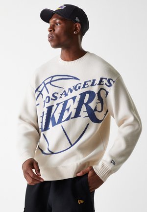 Cream sweater with a large navy "Los Angeles Lakers" graphic. Features a round neckline, ribbed cuffs, and textured knit. Worn with dark shorts.