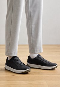 Dark gray sneakers with a textured upper, lace-up design, and a cushioned, light gray midsole. Worn with light gray tailored pants and white socks.