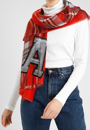 Écharpe à carreaux rouges avec une grande lettre grise "A" drapée sur un col roulé blanc, accompagnée d'un jean bleu taille haute porté par une personne debout.