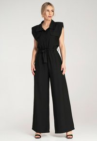Czarny kombinezon z poduszkami na ramionach, z zapięciem na guziki z przodu oraz wiązaniem w pasie. Szerokie, rozszerzające się nogawki i dwie przednie kieszenie. Gładka faktura materiału.