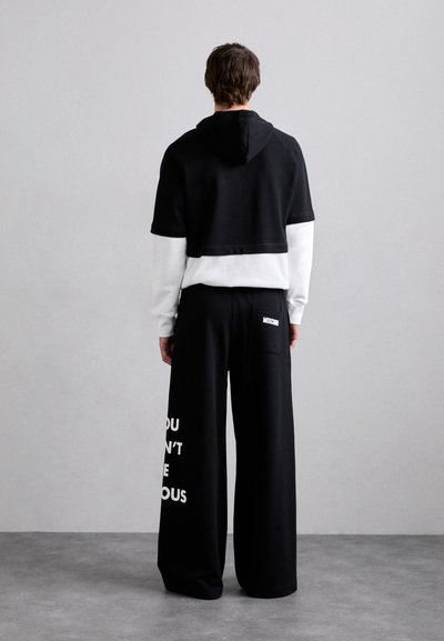 Čierna skrátená mikina s kapucňou vrstvená cez bielu košeľu s dlhými rukávmi, v kombinácii so širokými čiernymi nohavicami s bielym textom. Hladká tkanina, uvoľnený strih.