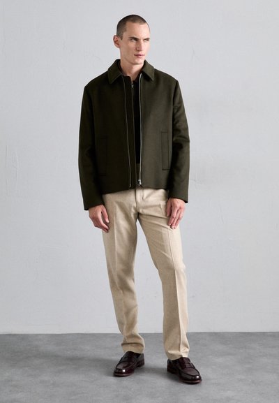 Olive green jacket with a zip closure, two front pockets, paired with beige pants and dark brown shoes, on a person standing against a neutral wall.