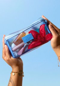 Mains tenant une trousse de maquillage transparente bleue contenant des produits cosmétiques et une fleur rouge, sur un fond bleu clair.