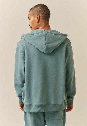 Mężczyzna stojący tyłem, ubrany w turkusową bluzę z kapturem i pasujące spodnie na jednolitym beżowym tle.