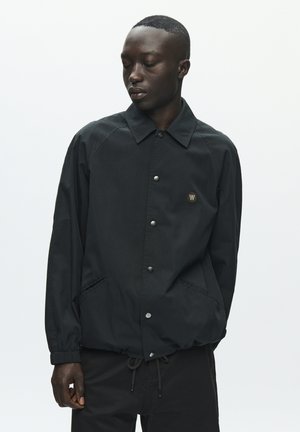 Noor mees, kes kannab musta nööbitavat jopet elastsete mansettide ja paelaga taljega, vaatab alla valgel taustal.