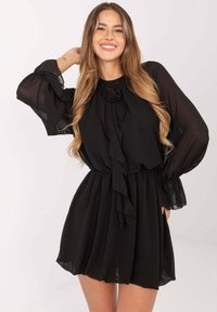 Czarna sukienka z przejrzystymi długimi rękawami, marszczonym dekoltem i wiązanym akcentem. Gładka tkanina; dopasowana w talii, rozszerzana na spódnicy.