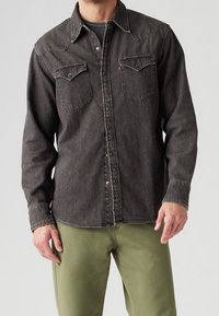 Uomo che indossa una camicia di jeans nera sbiadita con bottoni e tasche sul petto, insieme a pantaloni verdi oliva, in piedi contro uno sfondo grigio semplice.