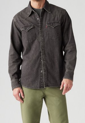Uomo che indossa una camicia di jeans nera sbiadita con bottoni e tasche sul petto, insieme a pantaloni verdi oliva, in piedi contro uno sfondo grigio semplice.