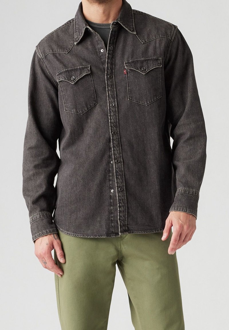 Uomo che indossa una camicia di jeans nera sbiadita con bottoni e tasche sul petto, insieme a pantaloni verdi oliva, in piedi contro uno sfondo grigio semplice.