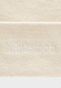 Kermavärinen neulekangas, jossa on kohopainettu, brodeerattu "lululemon" -logo samalla värillä keskellä. Pehmeä rakenne, ribatut reunat.