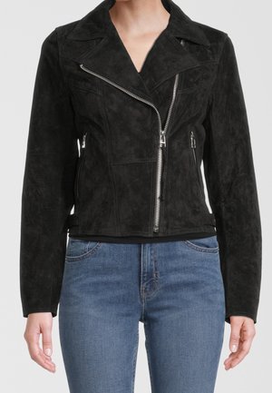 Kvinde iført en tætsiddende sort ruskind motorcykeljakke med sølv lynlåse og blå jeans, vist fra nakke til hofter mod en ensfarvet baggrund.
