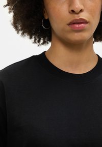 Juoda medvilnės džemperis su apvalia, supinta apykakle. Audinys atrodo minkštas, su lygia tekstūra, papildytu mažais žiediniais auskarais.