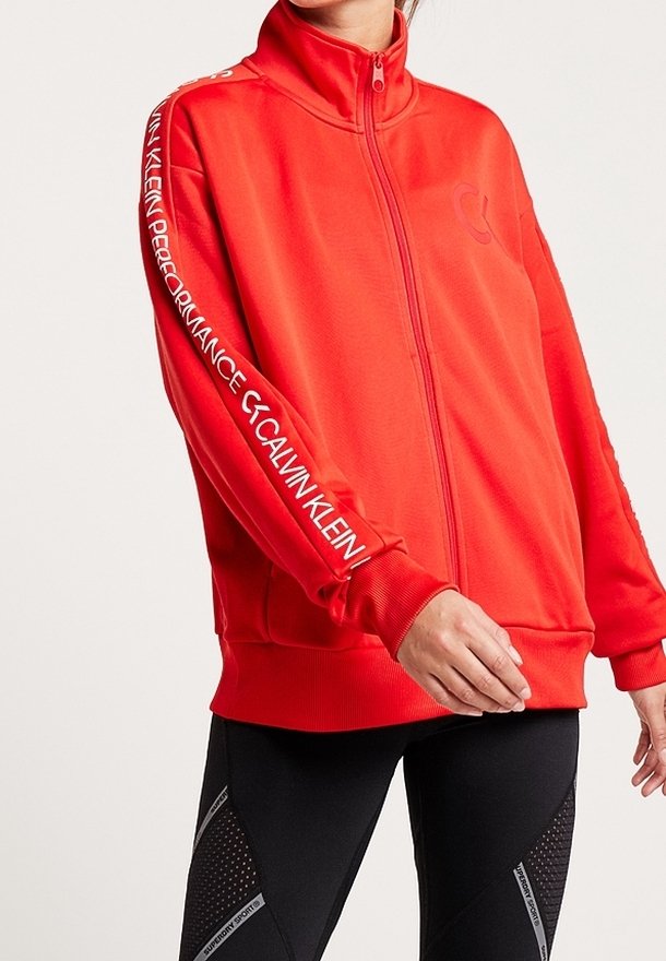 Rød zip-up jakke lavet af letvægtsstof, med høj krave og logo accenter på ærmerne. Glat tekstur, tætsiddende design.