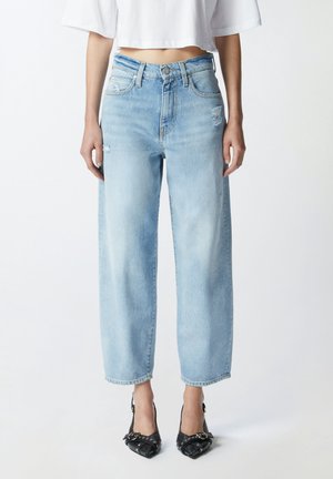 Vrouw draagt lichtblauwe jeans met hoge taille en rechte pijpen met lichte slijtage en zwarte puntige hakken, gecombineerd met een witte cropped top.