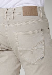 Närbild av beige denimjeansficka med sömdetaljer och lädermärke på midjebandet.