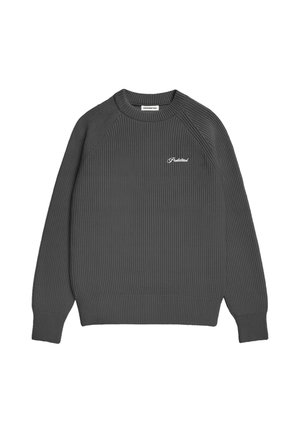 Homme portant un pull noir côtelé par-dessus une chemise à carreaux noir et blanc, un pantalon gris et un bonnet d’hiver en peau lainée noir et beige.
