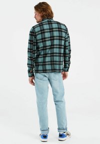Groen- en zwartgeruite flanellen shirt gedragen over lichtblauwe jeans. Het shirt heeft knoopmanchetten en een casual, ontspannen pasvorm.