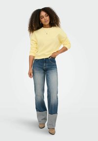 Femme aux cheveux bouclés portant un sweat-shirt jaune, un jean bleu avec les revers repliés et des chaussures pointues beige, debout avec une main sur la hanche.