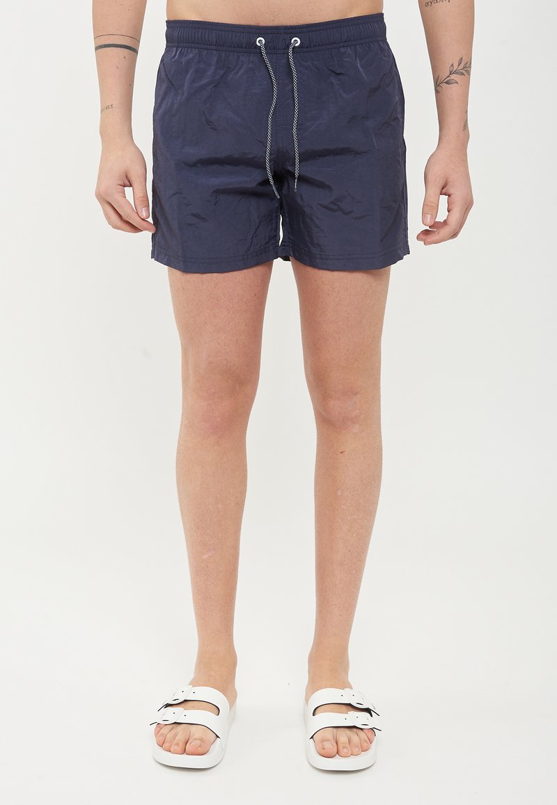Pantaloni da nuoto blu navy realizzati in materiale leggero, con vita elasticizzata, chiusura con cordoncino e orlo dritto. Indossati con sandali bianchi.